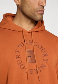 Orange hoodie lavet af blødt stof, med et cirkulært Nike-logo broderet i mørk lilla, med snøre-lukning og kengurulomme.