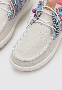 Las zapatillas de lona gris claro presentan acentos vibrantes con un patrón en azul, rojo y negro, con suelas redondeadas y costuras decorativas.