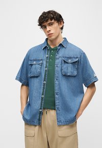 Νεαρός άνδρας με γυαλιά με διαφανή πλαίσια, μπλε κοντομάνικο τζιν πουκάμισο πάνω από πράσινο πουκάμισο με κουμπιά και μπεζ παντελόνι cargo, με τα χέρια στις τσέπες.