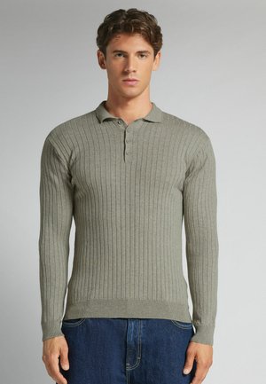 Junger Mann trägt einen hellgrauen, gerippten Polo-Pullover mit langen Ärmeln und blaue Jeans, steht vor einem einfarbigen Hintergrund.