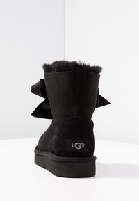 Černé sametové boty ve tvaru se zaoblenou špičkou, se světlou kožešinovou manžetou a velkou saténovou mašlí po straně. Logo UGG na patě.
