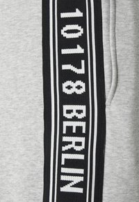 Grijze sweatshirt met een opvallende zwarte band met witte gebreide tekst "1017 BERLIN," omrand door twee witte strepen. Zachte textuur.