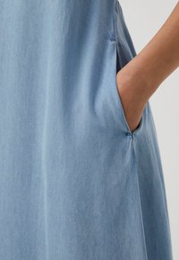 Rochie din denim de culoare albastru deschis, evidențiind o textură netedă, detalii cu buzunare laterale și cusături subtile pe cusături pentru un aspect sofisticat.