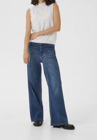 Jeans in denim scuro a gamba larga, dotati di cinque tasche e con una vestibilità rilassata. Abbinati a un top senza maniche grigio chiaro. Stivali neri alla caviglia.