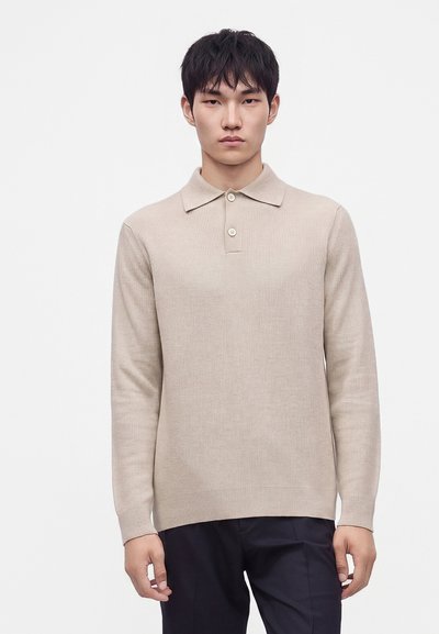 Jeune homme aux cheveux noirs portant un polo en maille beige à manches longues et un pantalon foncé, debout devant un fond blanc uni.