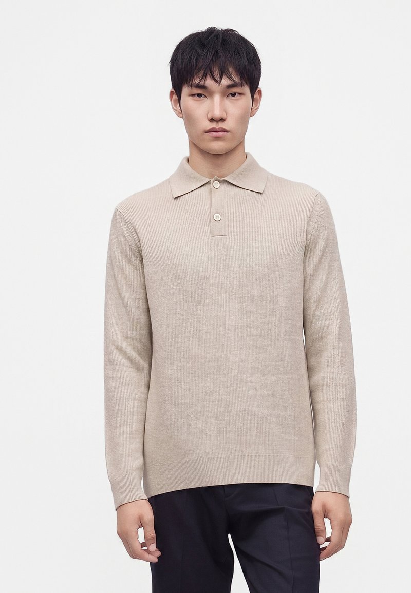 Jeune homme aux cheveux noirs portant un polo en maille beige à manches longues et un pantalon foncé, debout devant un fond blanc uni.