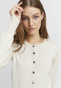 Cardigan bej tricotat cu decolteu rotund, șapte butoane negre pe față și manșete cu model in relief. Textură netedă cu o siluetă ajustată.
