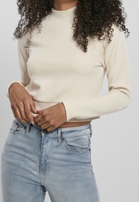 Top de manga larga beige claro con cuello alto falso, hecho de tela suave, con un diseño recortado y silueta ajustada. Combinado con jeans azules.