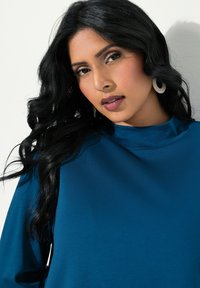 Teal långärmad topp med hög hals. Slät textur med minimala sömnadsdetaljer. Modellen har långt, vågigt mörkt hår och accentuerande örhängen.