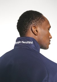 Tamsiai mėlynas sportinis švarkas su aukštu apykakle, tekstūruotas audinys ir baltas "Sergio Tacchini" logotipas apykaklėje. Detalus galinės pusės vaizdas.