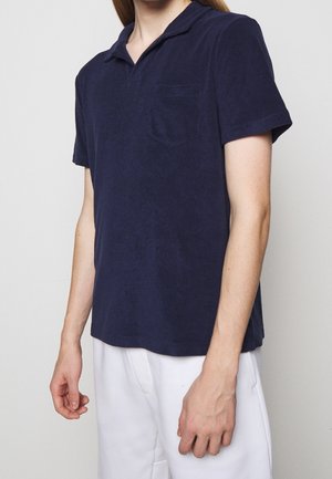 Personne portant un polo bleu marine à manches courtes avec une poche poitrine et un pantalon blanc, debout devant un fond gris uni.