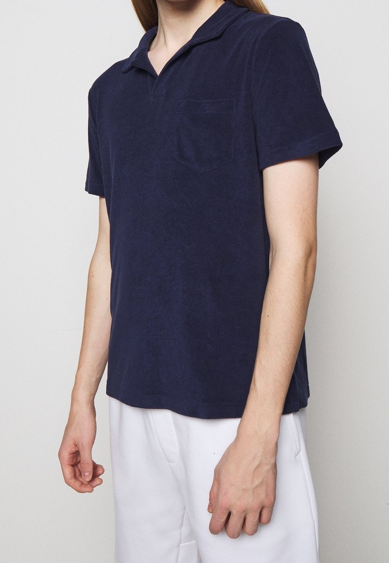 Personne portant un polo bleu marine à manches courtes avec une poche poitrine et un pantalon blanc, debout devant un fond gris uni.