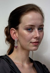 Young woman with blue eyes and freckles wearing long silver earrings with red and clear gemstones, black top, and hair tied back.