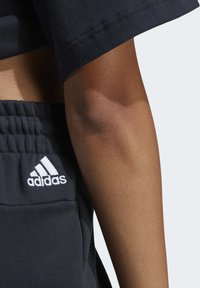 Čierny bavlnený crop top s krátkymi rukávmi, skombinovaný s tmavými teplákmi s bielym logom Adidas na vrecku. Hladká, štruktúrovaná tkanina.