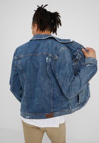 Person with dreadlocks wearing a blue denim jacket over a white shirt and beige pants, viewed from the back against a plain background.