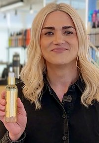 Bottiglia di vetro trasparente riempita con liquido dorato, con un tappo e un'etichetta dorati. Persona che la tiene, indossando una camicia nera a bottoni.