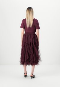 Donna in piedi con la schiena rivolta, che indossa un top corto bordeaux e una gonna midi in tulle a strati, con scarpe nere con tacco basso, su sfondo bianco.