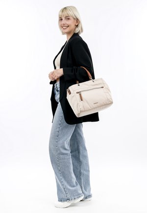 Sac à main beige avec poignée en cuir marron, poche zippée, forme rectangulaire, texture de tissu douce et détails de couture bien visibles.