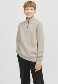 Ljusbeige zip-up sweatshirt med hög krage, ribbade ärmslut och nederkant. Bärs med svarta byxor. Slät textur, minimalistisk design.
