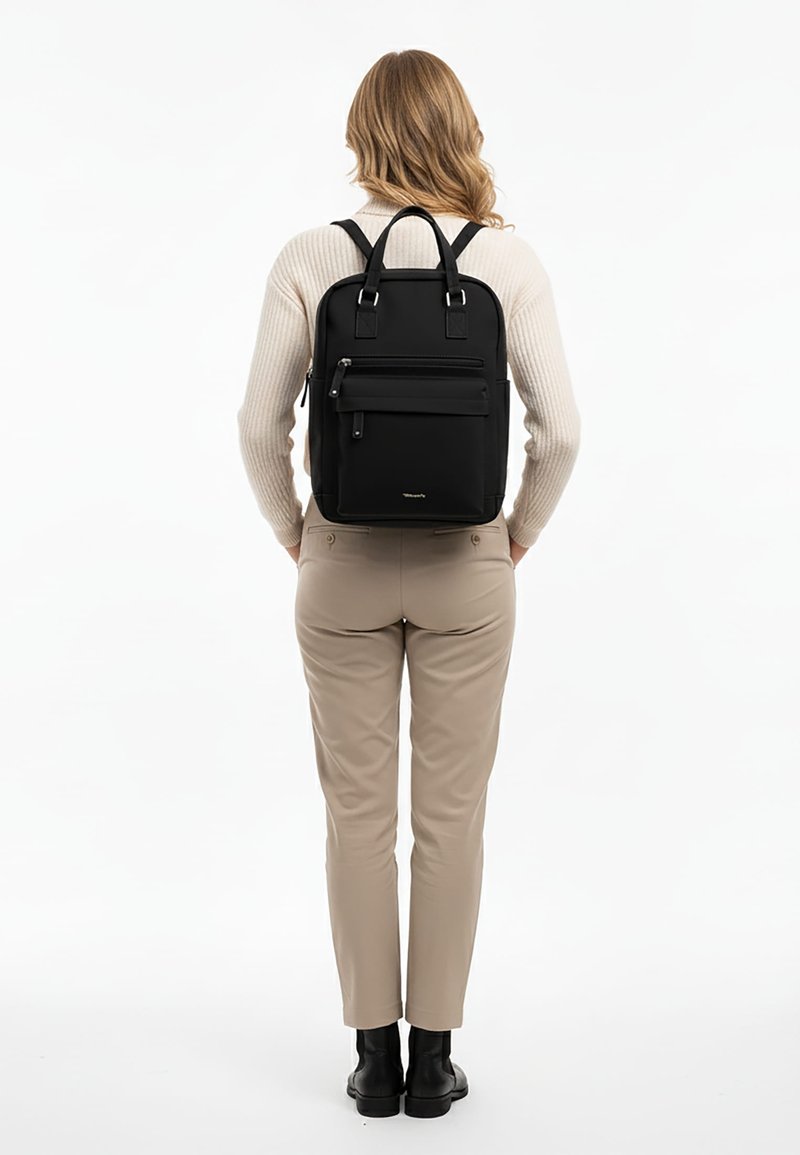 Vrouw met lang golvend haar, beige broek, crèmekleurige trui, zwarte laarzen en een zwarte rugzak met ritsvakken aan de voorkant, staand tegen een witte achtergrond.