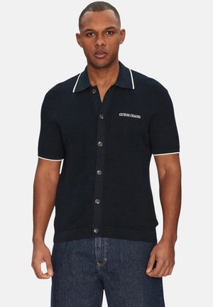 Navyfarbendes, strukturiertes Hemd mit Knopfleiste, kragen und weißen Akzenten sowie gesticktem "GUESS JEANS"-Logo, kombiniert mit dunkler Jeans.