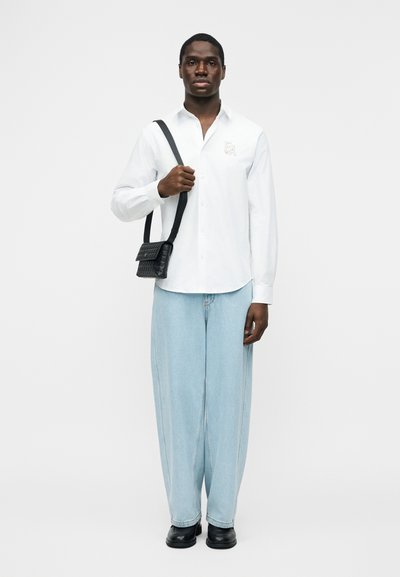 Άνδρας που φοράει λευκό πουκάμισο με κουμπιά, ανοιχτό μπλε τζιν παντελόνι φαρδύ, μαύρα παπούτσια, κρατώντας μικρή μαύρη τσάντα χιαστί με ιμάντα υφαντής υφής.