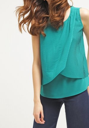 Femme portant un chemisier sans manches plissé de couleur bleu sarcelle associé à un pantalon bleu foncé, avec des cheveux bruns ondulés partiellement visibles.