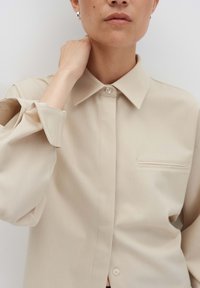 Persona con una camicia beige con bottoni, colletto ampio e maniche arrotolate, che tocca il collo, su uno sfondo chiaro e semplice.