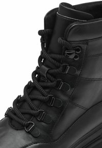 Botte en cuir noir avec un design haut de cheville, des lacets épais et texturés, et des œillets en métal à l'avant avec une finition lisse.