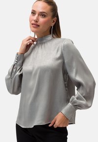Blusa di raso argento con collo alto, maniche a sbuffo lunghe con dettagli a bottoni e orlo leggermente curvo. Tessuto liscio con un effetto lucido.