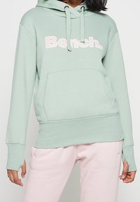 Hoodie verde menta con tasca anteriore, vestibilità ampia e cappuccio con cordino. Presenta il testo bianco "Bench." sul petto. Tessuto morbido, polsini a coste.