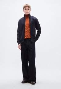 Μπλε μπουφάν με φερμουάρ πάνω από πορτοκαλί ζιβάγκο, συνδυασμένο με παντελόνι από σκούρο τζιν φαρδύ και καφέ παπούτσια· απαλές υφές και καθαρές γραμμές.