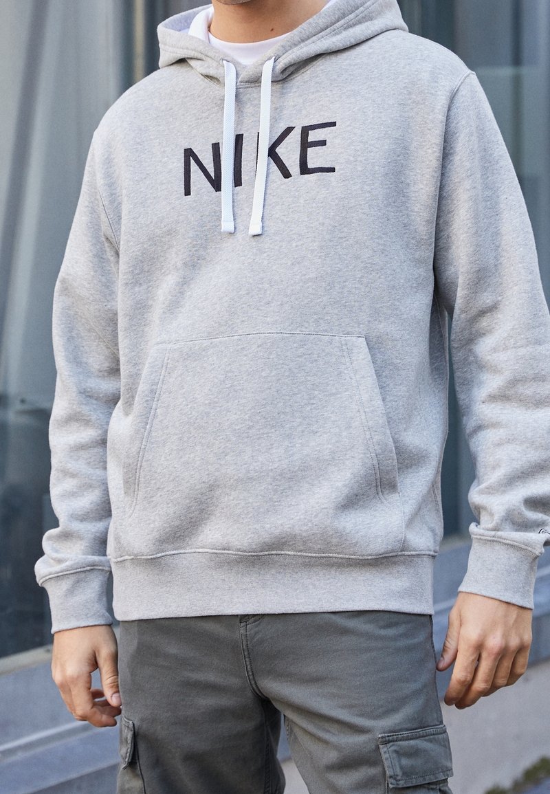 Šedá mikina s klokáním kapsou, bílými šňůrkami a černým logem "NIKE" na přední straně. Měkká textura a pohodlný střih.