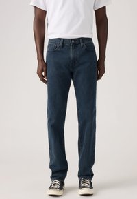 Jeans de ganga azul escuro com design de perna reta, estilo com cinco bolsos e fecho com botão metálico. Usados com sapatilhas pretas e uma t-shirt branca.