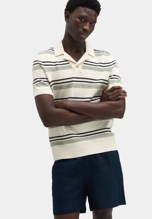 Man with crossed arms wearing a short-sleeve collared shirt with horizontal stripes and dark shorts against a plain background.