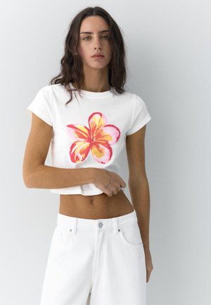 Žena s dugom tamnom kosom u bijeloj skraćenoj majici s velikim ružičasto-žutim cvjetnim uzorkom i bijelim hlačama na jednobojnoj pozadini.