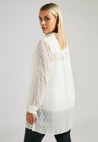 Camicia bianca a fantasia con maniche lunghe e collo abbottonato, con una texture semi-trasparente e un orlo posteriore leggermente più lungo.