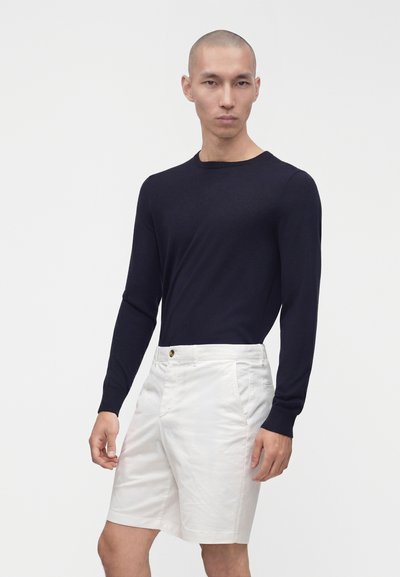 Homme portant une chemise noire à manches longues et un short blanc jusqu'aux genoux, debout devant un fond gris clair uni.