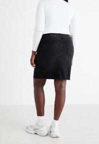 Osoba oblečená v bílém dlouhém rukávu, černé denimové sukni, bílých ponožkách a bílých teniskách, stojící proti jednoduchému světlému pozadí.