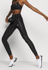 Leggings sportivi neri con finitura lucida, con logo e strisce bianche PUMA. Abbinati a sneaker bianche e a una borsa in vita nera.