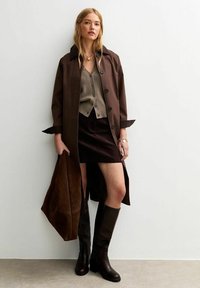 Abrigo trench marrón sobre un suéter gris de botones, combinado con una falda de pana oscura y botas marrones hasta la rodilla, sosteniendo un bolso tote de ante marrón.