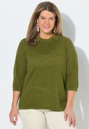 Lachende vrouw met lichtbruin haar draagt een olijfgroene geribbelde trui met driekwart mouwen en creme broek tegen een witte achtergrond.