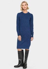 Abito blu lavorato a maglia con maniche lunghe, scollatura rotonda e orlo a coste. Abbinato a stivali neri in pelle fino al ginocchio. Sfondo neutro.