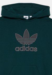 Temnozeleni kapuča s sweatshirtom, ki prikazuje velik, prišit Adidasov trefoil logotip v sivi barvi s tremi vodoravnimi črtami. Nežen teksturiran material.