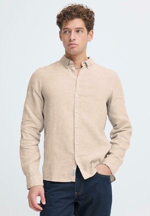 Lyse beige linnedragt med knaplukket krave, lange ærmer og en lomme. Bruges over mørke jeans med synlige syninger.