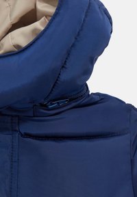 Primo piano di una giacca a vento blu navy con cappuccio testurizzato, tessuto liscio e una cerniera blu al collo con un rivestimento interno di colore crema.