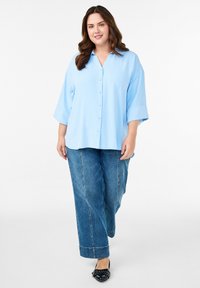 Woman with long brown hair walking, wearing a light blue buttoned blouse, wide-leg blue jeans, and black flats with a buckle.