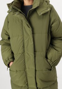 Person wearing an olive green padded winter jacket with hands in side pockets, partially zipped over a black shirt.