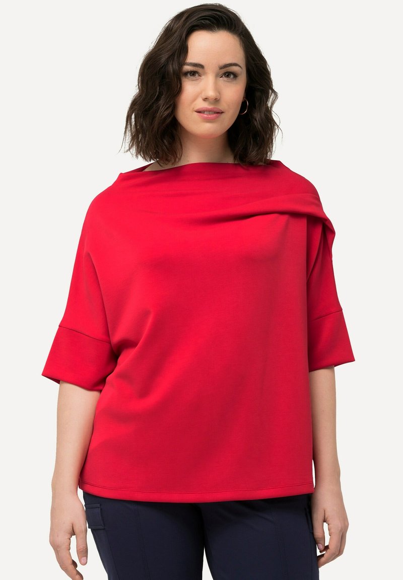 Maglia rossa con un ampio scollo drappeggiato e maniche corte. Texture liscia e vestibilità leggermente ampia. Nessun motivo visibile.