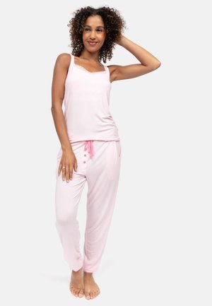 Vrouw met krullend haar, gekleed in een lichtroze mouwloos topje en bijpassende pyjamabroek, staat blootsvoets met een hand op haar hoofd.